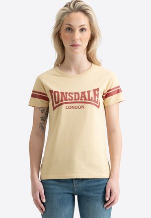 Joven mujer con pelo rubio que lleva una camiseta beige de Lonsdale London con rayas rojas en las mangas y vaqueros azules, de pie frente a un fondo blanco.