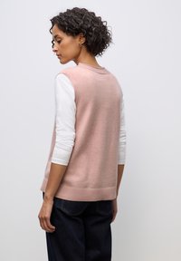 Světle růžový svetřík bez rukávů s vystuženým lemem, nošený přes bílou dlouhými rukávy. Má jednoduchý design a jemnou texturu.