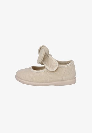 Zapato para niño pequeño de tela beige con punta redondeada, una correa de velcro y un nudo decorativo en la correa, mostrado de lado.
