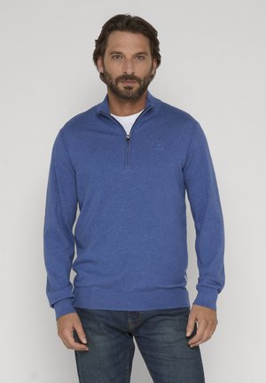 Mężczyzna z brodą w niebieskim swetrze na zamek błyskawiczny, na białej koszuli i niebieskich dżinsach, stojący na jasnym, jednolitym tle.