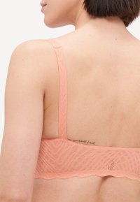 Sutiã de renda pêssego com padrão zigzag texturizado, alças ajustáveis e design de borda em scallop. Apresenta uma etiqueta logo em flat.