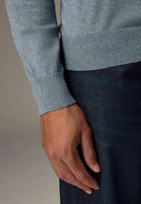 Helleblaue Pullover mit gerippten Bündchen, kombiniert mit dunkler Jeans. Nahaufnahme zeigt eine Hand, die auf dem Stoff mit glatter Textur ruht.