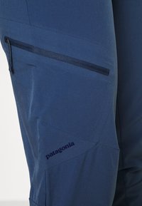Pantalons de plein air bleu marine en tissu texturé, dotés d'une poche zippée sur le côté et du logo Patagonia, conçus pour la durabilité et la fonctionnalité.