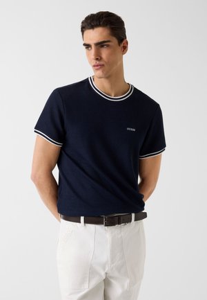 Uomo che indossa una camicia a maniche corte blu navy con rifiniture bianche e logo "GUESS", pantaloni bianchi e cintura marrone scuro, in piedi davanti a uno sfondo semplice.