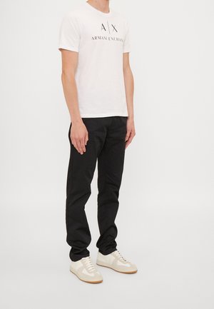 Hombre con camiseta blanca de Armani Exchange, pantalones negros y zapatillas blancas, posando frente a un fondo claro y sencillo.