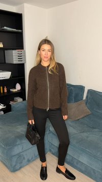 Cardigan zippé marron pelucheux, pantalon noir ajusté et mocassins noirs. Canapé texturé bleu sarcelle en arrière-plan avec une décoration neutre.