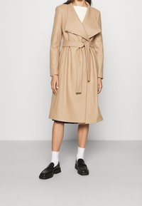 Beige uldblandet trenchcoat med en omslag design, lange ærmer og bælte i taljen. Har en bred revers og subtile detaljer i guldfarvet hardware.