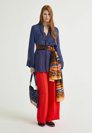 Jeune femme aux longs cheveux roux portant un chemisier bleu marine, un pantalon large rouge, une ceinture orange à motifs et tenant un sac à main bleu marine à franges.