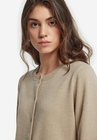 Beige kashmirtröja gjord av mjukt tyg, med rund halsringning, knappstängning och subtila ribbade detaljer längs kanterna.