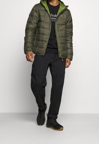 Chaleco de plumas verde con capucha, cierre frontal, combinado con un sudadero negro, pantalones oscuros y calzado negro con suelas de agarre texturizado.