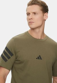 T-shirt in cotone verde oliva con collo a giro, caratterizzato da un design a tre righe nere sulla spalla sinistra e logo sul petto.