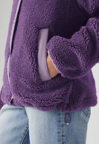 Lila polárdzseki texturált külsővel, világoslila belső béléssel, oldalzsebekkel és állítható alsó szegéllyel.