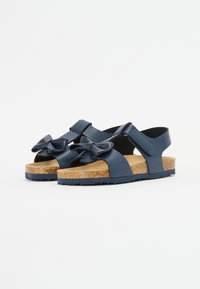 Mørkeblå sandaler med en teksturert korksåle, med brede stropper og en stor sløyfedetalj foran. Borrelås for enkel påkledning.