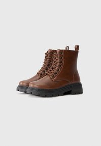 Botas de cuero marrón con cordones hasta el tobillo y suelas gruesas de goma negra sobre un fondo claro y liso.