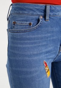 Blå denimjeans med en klassisk femficksdesign, med broderat läppstiftsmotiv på vänster lår.