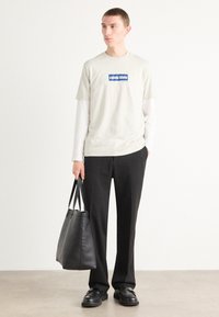 Lys grå t-skjorte med en blå logo, lagdelt over en hvit langermet skjorte. Sorte bukser og en svart lær totebag. Sorte sko.