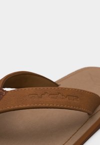 Barna bőr papucs, varrott pánttal, melyen márkajelzés dombornyomott, közeli felvételen világos, egyszerű háttéren.