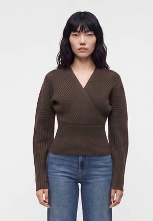 Femme aux cheveux noirs portant un pull cache-cœur en maille côtelée marron et un jean bleu sur un fond blanc uni.