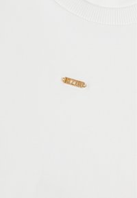 Biała bawełniana koszulka z teksturowanym złotym metalowym znaczkiem z napisem "BIZOU" z przodu, okrągły dekolt z prążkowanym wykończeniem.