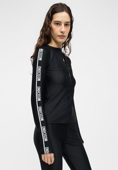 Γυναίκα με μακριά σγουρά μαλλιά φοράει μαύρο μπουφάν με φερμουάρ και παντελόνι· τα μανίκια του μπουφάν έχουν λευκές ρίγες με το κείμενο "MOSCHINO".