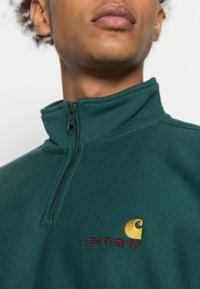 Pullover verde com zip até ao peito, feito de tecido suave. Apresenta um logotipo bordado em dourado e bordô no peito e um colarinho estruturado.