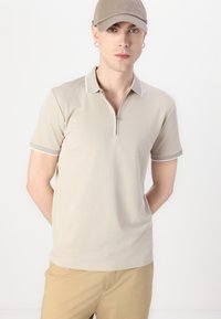 Beige polo-shirt med kort erm, glidelås krage og kontrasterende hvite sting ved kragen og ermene.