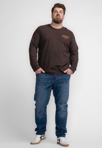 Donkerbruine lange mouwen shirt met een geborduurd oranje logo, gecombineerd met blauwe jeans en witte sneakers met zwarte accenten.