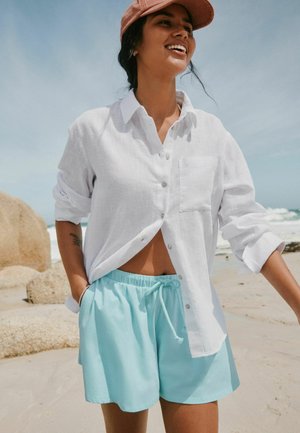 Femeie zâmbitoare pe o plajă, purtând o cămașă albă cu nasturi, pantaloni scurți bleu cu șnur și o șapcă roz, cu mâinile în buzunare.