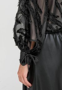 Top nero textured con tessuto trasparente, caratterizzato da strisce nere pelose e polsini legati. Indossato sopra una gonna in pelle nera liscia.