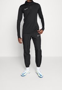 Black athletic pullover with a high collar and quarter zipper, featuring white side stripes. Matching black tapered pants, both with Nike logos.