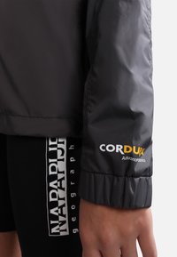 Grå pufferjakke med ribbet manchet. Logoen "CORDURA" i gul og hvid på ærmet. Glat, skinnende stof og afslappet pasform.