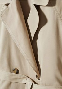 Détail d'un trench beige avec gros plan sur le col, les coutures et deux grands boutons ronds. Tissu lisse avec une ceinture nouée à la taille.