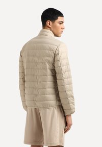 Giacca a vento beige con design trapuntato, colletto alto e materiale leggero, abbinata a pantaloni corti beige, mostrata da dietro su uno sfondo bianco.
