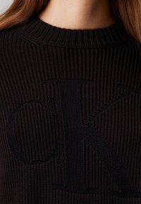 Černý pletený svetr s žebrovanou texturou a výrazným černým logem "OK". Výstřih je klasického žebrovaného střihu.