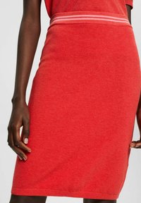 Punainen neulepulloverihame, jossa on teksturoitu pinta. Ominaisuuksiin kuuluu leveä vyötärönauha, jossa on kaksi vaaleanpunaista raitaa, liukuvasti istuva muotoilu ja polvipituinen leikkaus.