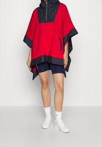 Röd poncho med en vadderad svart övre del, huva, sidofickor och marinblå detaljer. Kombinerad med marinblå shorts och vita sneakers.
