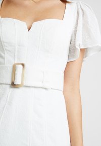 Biała sukienka z teksturowanej tkaniny, kwadratowym dekoltem, bufiastymi rękawami i szerokim paskiem z prostokątną kremową klamrą. Widoczne szczegółowe przeszycia.