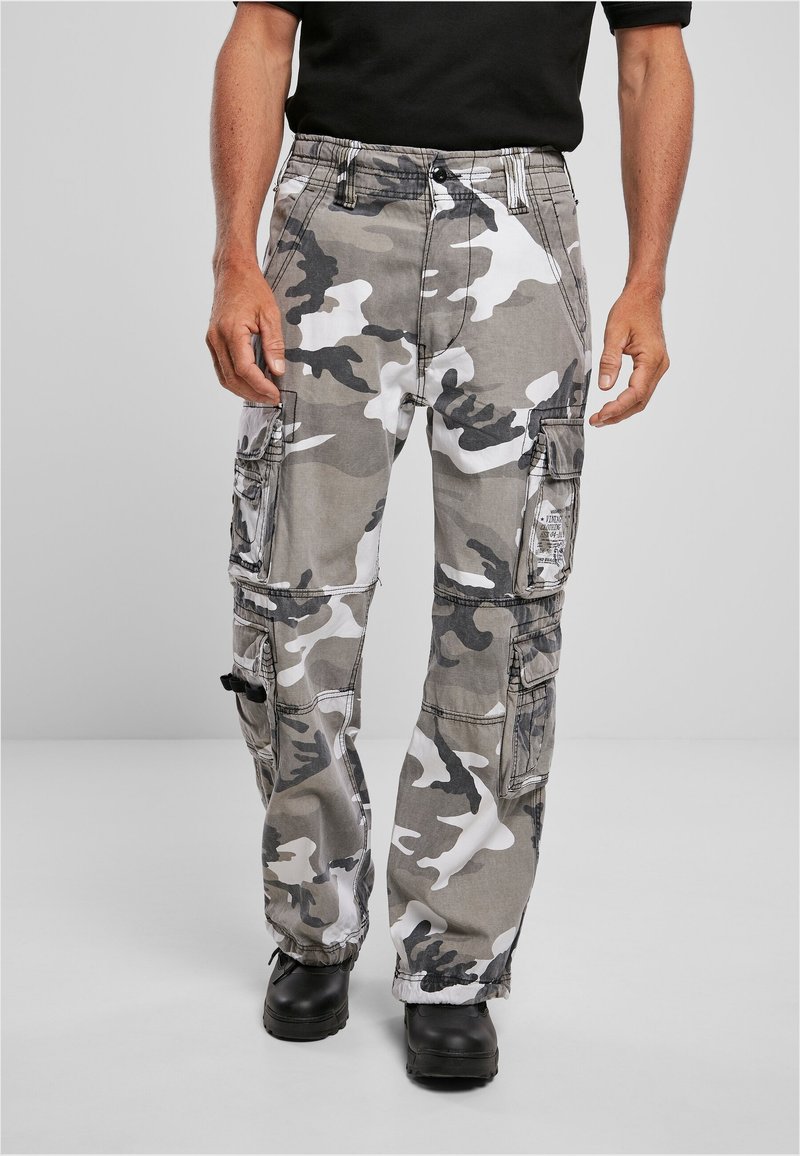 Homme portant un pantalon cargo camouflage gris, blanc et noir avec plusieurs poches et des chaussures noires, debout devant un fond uni.