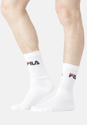 Jambes portant des chaussettes blanches côtelées à tige moyenne avec le logo FILA en noir et rouge, un pied à plat et l'autre sur la pointe contre un fond blanc.