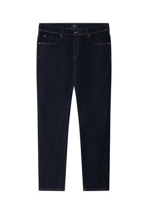 Homme portant un blazer blanc, une chemise rose clair, un jean foncé, une ceinture marron et des mocassins marron, debout devant un fond gris uni.