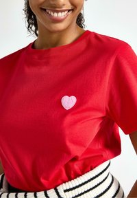 Czerwona bawełniana koszulka z małą różową łatą w kształcie serca na piersi, w połączeniu z poziomo paskowanym czarno-białym pasem.
