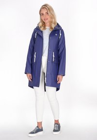 Marinblå vattentät regnjacka med huva, dragkedja och snörning. Har två framfickor och vita detaljer. Bärs över en randig skjorta och vita byxor.