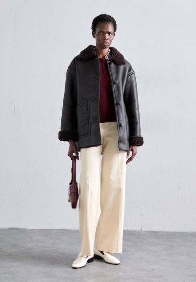 Crna kožna jakna s ovratnikom od smeđeg umjetnog krzna, uparen s bordo pletenim topom i kremastim širokim hlačama. Model drži tamnocrvenu torbu.