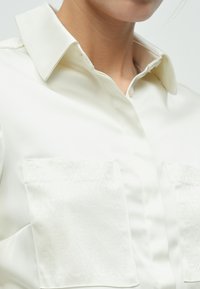 Λευκό μπλουζάκι από σατέν με μυτερό γιακά, που διαθέτει δύο μπροστινές τσέπες με ανάγλυφα διακοσμητικά σε σχήμα διαμαντιού. Λείο υφή και εφαρμοστό σχέδιο.