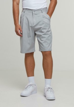 Man wearing light gray pleated shorts, white crew socks, and white sneakers, standing with one hand in pocket on plain background.