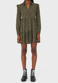 Zwart-groen patroon jurk met lange mouwen, een kraag, ruches als accent en een uitlopende zoom, gecombineerd met zwarte veter-enkellaarzen.