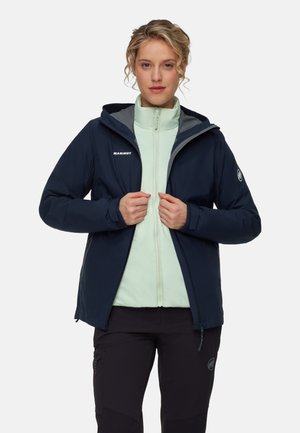 Vrouw in een marineblauwe hoodie over een lichtgroene ritsjack en zwarte broek, staand tegen een witte achtergrond.
