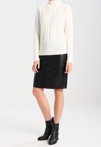 Cremfarbener Strickpullover mit einem Zopfmuster, kombiniert mit einem schwarzen Leder-Bleistiftrock. Schwarze Knöchelboots mit einem Reißverschluss-Detail runden den Look ab.