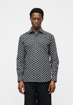 Homme portant une chemise à manches longues à motifs noirs et blancs avec des boutons et un pantalon noir, debout devant un fond clair uni.