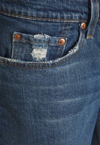 Siniset farkut, joissa on kulunut tasku repsottavilla reunoilla ja kuparinvärisillä metalliosilla. Kangas näyttää kuluneelta ja haalistuneelta.